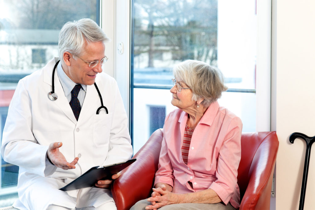 Male doctor in consultation with a senior female patient sitting having a discussion and offering a detailed explanation as they chat in front of a window in the hospital